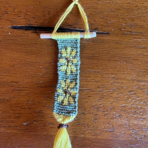 May include: A small, hand-woven wall hanging with a yellow tassel. The hanging features a green and yellow geometric pattern with a floral design.