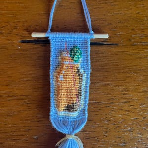 May include: A small blue and orange woven wall hanging with a tassel. The hanging features a cartoon image of a person holding a green drink.