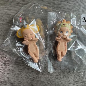 May include: Two small figurines in clear plastic packaging. One figurine is a light tan color with yellow ears and a small hat. The other figurine is light tan with a lion's mane and a small hat. A white sticker with the number 3 is visible.