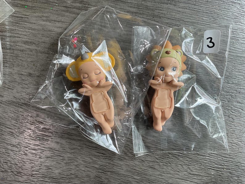 May include: Two small figurines in clear plastic packaging. One figurine is a light tan color with yellow ears and a small hat. The other figurine is light tan with a lion's mane and a small hat. A white sticker with the number 3 is visible.