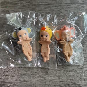 May include: Three small figurines in clear plastic packaging. The figurines are nude-colored with painted faces and animal-themed hats: a gray koala, a yellow duck, and an orange elephant. The figurines are in a standing pose with arms outstretched.