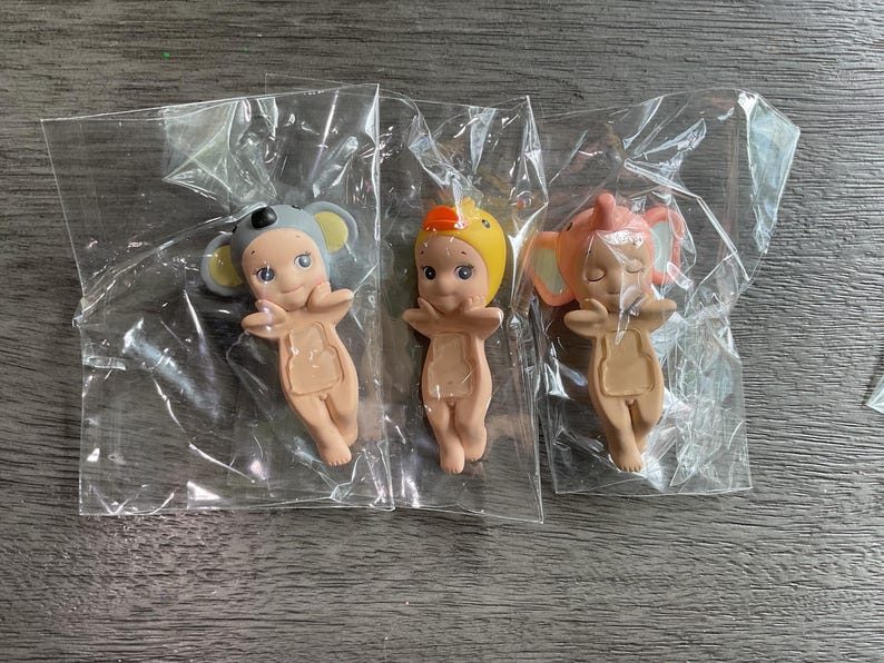 May include: Three small figurines in clear plastic packaging. The figurines are nude-colored with painted faces and animal-themed hats: a gray koala, a yellow duck, and an orange elephant. The figurines are in a standing pose with arms outstretched.