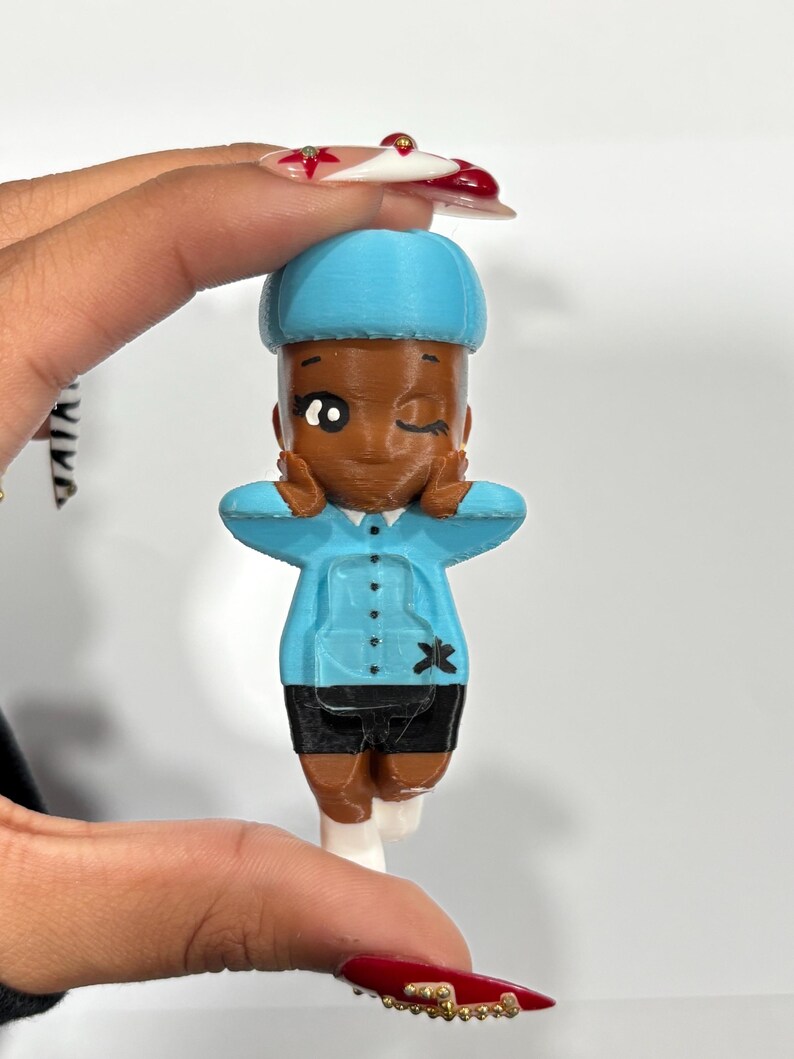 May include: A 3D-printed figurine of a person with brown skin, wearing a light blue uniform with black shorts and white shoes. The figurine is winking and making a peace sign with its hands. The figurine has a blue hat.