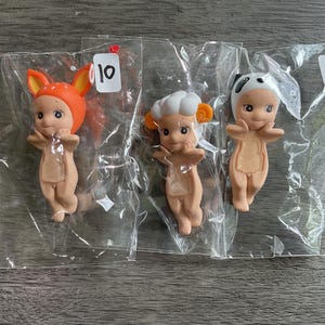 May include: Three small figurines in clear plastic bags. One figurine wears an orange fox hat, another a chef's hat, and the third a panda hat. Each figurine has a light brown body and is in a standing pose. The figurines are numbered 9 and 10.