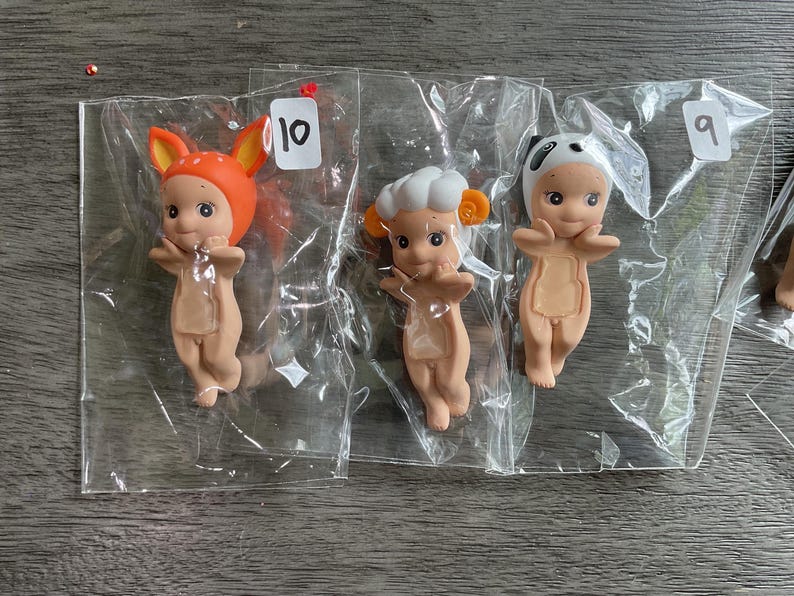 May include: Three small figurines in clear plastic bags. One figurine wears an orange fox hat, another a chef's hat, and the third a panda hat. Each figurine has a light brown body and is in a standing pose. The figurines are numbered 9 and 10.