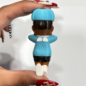 May include: A 3D-printed figurine of a person, viewed from the back. The figure wears a light blue hat, jacket, and white shoes. The jacket has small white wings on the shoulders. The figure has dark hair and skin, and black shorts.