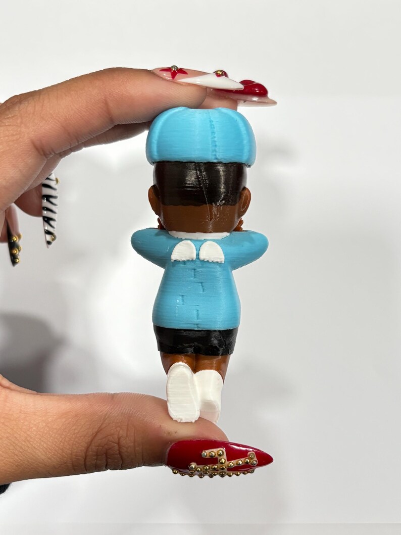 May include: A 3D-printed figurine of a person, viewed from the back. The figure wears a light blue hat, jacket, and white shoes. The jacket has small white wings on the shoulders. The figure has dark hair and skin, and black shorts.