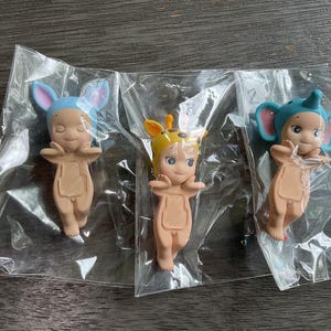 May include: Three small figurines in clear plastic packaging. Each figurine is a nude-colored baby with a different animal-themed hat: a blue bunny, a giraffe, and a blue elephant. The figurines have closed eyes and outstretched arms.