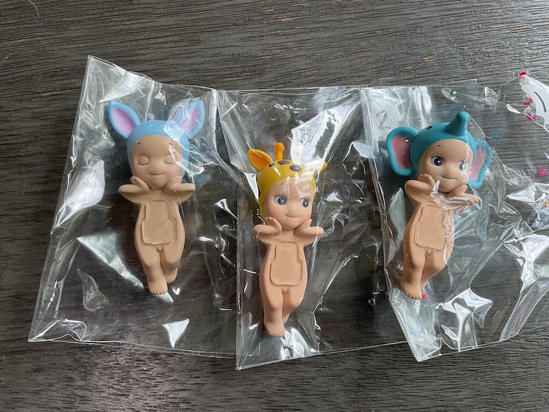 May include: Three small figurines in clear plastic packaging. Each figurine is a nude-colored baby with a different animal-themed hat: a blue bunny, a giraffe, and a blue elephant. The figurines have closed eyes and outstretched arms.