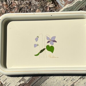 Puede incluir: Una bandeja rectangular de metal color crema con esquinas redondeadas y un borde elevado. El centro presenta una ilustración gráfica de una flor de violeta morada con hojas verdes y tallos delgados.
