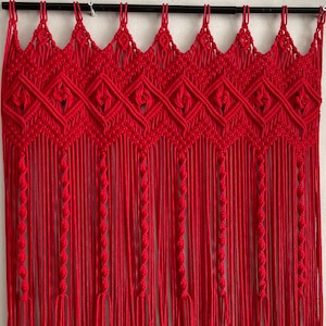 May include: A red macrame wall hanging with a geometric pattern and fringe. The hanging is made of thick cotton cord and has a simple, modern design.