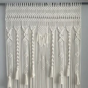 Könnte beinhalten: Ein weißer Makramee-Wandbehang mit geometrischem Muster und Quasten. Der Behang ist aus Baumwollschnur gefertigt und etwa 90 cm breit und 120 cm hoch.