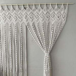 Puede incluir: Colgante de pared de macramé blanco con un patrón geométrico. El colgante está hecho de cordón de algodón y tiene una franja en la parte inferior.