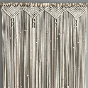 May include: A white macrame curtain with a wood dowel rod at the top. The curtain is made of a series of knotted cords that create a decorative pattern.