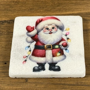 May include: A white ceramic coaster with a festive image of Santa Claus holding a string of colorful lights. Santa is wearing a red suit with white trim and a white beard. He is smiling and waving.