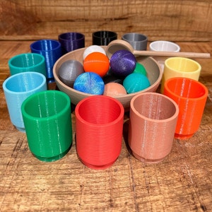 Könnte beinhalten: Ein Set aus bunten Holzbechern und -kugeln, perfekt für ein Spiel zum Stapeln oder Sortieren. Die Becher sind in verschiedenen Blautönen, Grün, Rot und Braun, während die Kugeln eine Mischung aus Blau, Grün, Rot und Weiß sind.