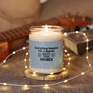 May include: A white candle with a blue label that reads "Everything Happens For a Reason And It's Called PHYSICS". The label has two atomic symbols on it. The candle is sitting on a wooden coaster.