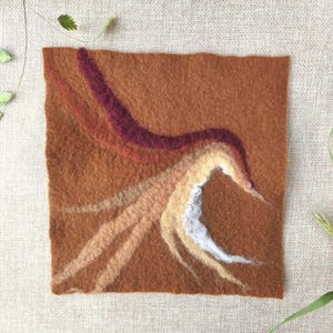 May include: A square, felted textile art piece in shades of brown, burgundy, and cream. The design features flowing, abstract shapes resembling a bird or a flame. The artwork is set against a neutral, textured background with sprigs of greenery.