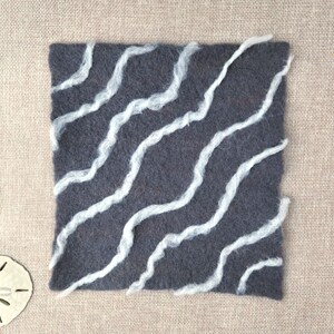 May include: A square dark gray felt mat featuring diagonal, wavy white wool fibers across the surface. The mat is accompanied by a light-colored sand dollar and a small, light-colored starfish placed near the corners of the square felt piece.