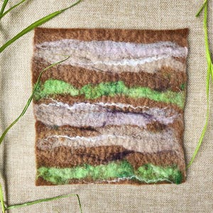May include: A square, felted textile with horizontal bands of brown, green, white, and gray. The textured surface resembles a landscape. Green grass blades are in the foreground, adding a natural element to the composition.