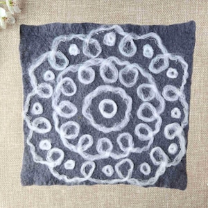 May include: A square, dark gray felted wool piece with a white, circular, floral-like design. The design is made of looped yarn. The piece is on a neutral-colored linen surface, with white flowers in the corners.