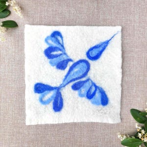 May include: A square, white felted wool piece with a blue bird design. The bird is depicted with stylized wings and tail, and a teardrop-shaped head. The design is in shades of blue. The background is a neutral linen fabric.