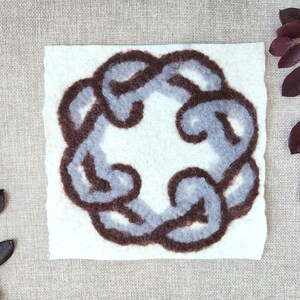 May include: A square, off-white felted artwork featuring a stylized, interlocking chain design in shades of gray and brown. The artwork is surrounded by dark red-purple leaves and stems, creating a natural, artistic composition.