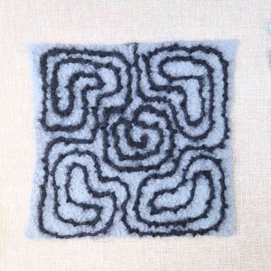 May include: A square, light blue felted textile with a dark gray, swirling, maze-like design. The textile is surrounded by a neutral background with a few small, colorful stones.