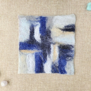 Könnte beinhalten: Quadratischer Filztextil mit einem abstrakten Design in Blau-, Weiß- und Schwarztönen. Das Kunstwerk hat ein strukturiertes Aussehen. Der Hintergrund ist ein neutraler beigefarbener Stoff. Kleine dekorative Steine und ein Seestern.