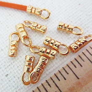 May include: Gold-colored crimp end findings with a loop for jewelry making. The findings have a textured, cylindrical design. An orange cord is attached to one of the findings. A ruler is in the background.