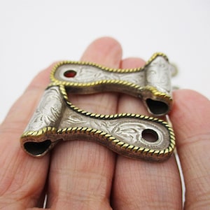 May include: Two silver-toned metal objects with intricate floral engravings and gold-colored trim. The objects have a curved shape with a hole at one end and a small red accent. The design suggests a vintage or handcrafted style.