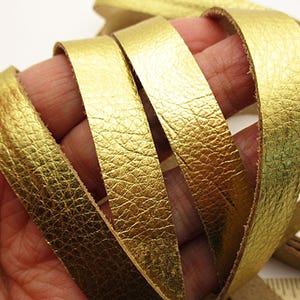 May include: A long, flexible strip of metallic gold leather with a visible textured grain surface. The leather strip is coiled in loops and positioned next to a ruler showing measurements in inches to indicate the width of the material.