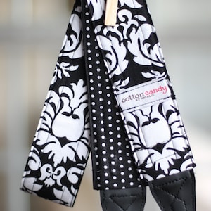 May include: Black and white camera strap with a polka dot pattern and a damask pattern. The strap has a black leather adjustable section and the text "cotton candy by Natalie" is printed on the strap.