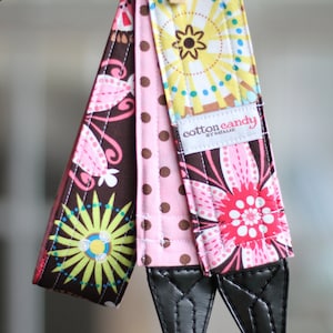 May include: A pink, brown, and blue camera strap with a floral pattern and polka dots. The strap has a black leather adjustable strap with a "cotton candy by Natalie" label.