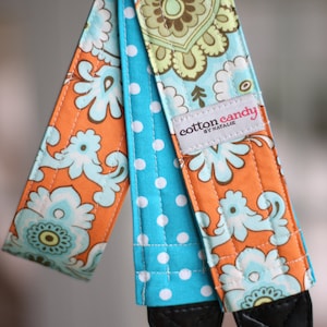 May include: A camera strap with a turquoise and white polka dot fabric, a turquoise and orange floral fabric, and a black leather strap. The strap has a label that reads "cotton candy by natalie".