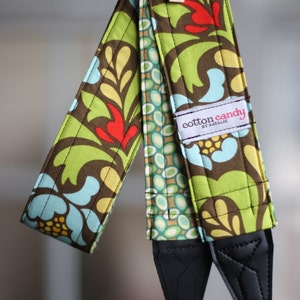 May include: A green, brown, and blue patterned camera strap with a black adjustable strap and a label that reads "cotton candy by Natalie".