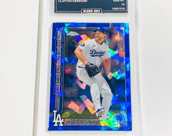 May include: A baseball trading card featuring Clayton Kershaw in a Dodgers uniform. The card is encased in a protective holder and has a blue, gem-like design. The text on the card reads "2025 Topps Sapphire" and "Clayton Kershaw."