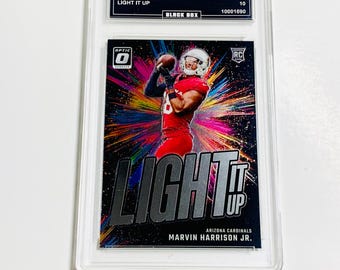 May include: A graded sports trading card featuring Marvin Harrison Jr. in a red Arizona Cardinals uniform. The card has the text "LIGHT IT UP" and "2024 Donruss Optic". The card is encased in a clear protective holder.