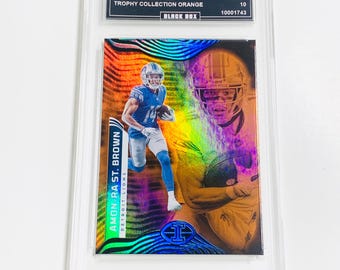 May include: A trading card featuring Amon-Ra St. Brown of the Detroit Lions. The card is from the 2022 Illusions Trophy Collection Orange series, graded GEM MT 10. The card has a holographic design with a player image.
