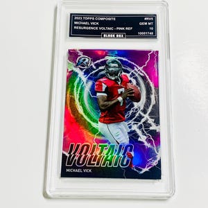 May include: A Michael Vick football trading card in a protective case. The card features a player in a red uniform against a pink and purple background with lightning effects. Text includes "VOLTAIC" and "2023 TOPPS COMPOSITE".