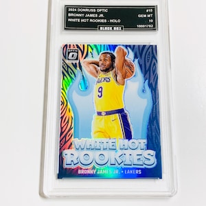 May include: A 2024 Donruss Optic Bronny James Jr. White Hot Rookies - Holo basketball card, encased in a protective holder. The card features a player in a yellow Lakers jersey, with a blue and white flame design.