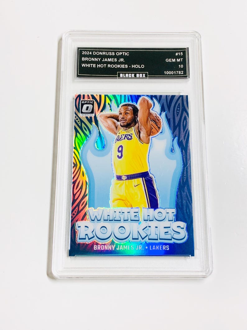 May include: A 2024 Donruss Optic Bronny James Jr. White Hot Rookies - Holo basketball card, encased in a protective holder. The card features a player in a yellow Lakers jersey, with a blue and white flame design.