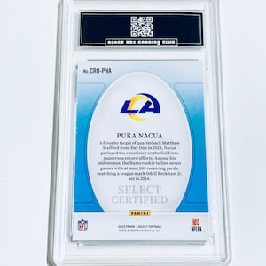 May include: A certified Puka Nacua football card from the 2023 Panini Select series, encased in a protective holder. The card features the LA Rams logo and details about Nacua's performance. The holder is labeled by the Black Box Grading Club.