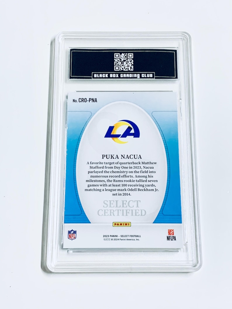 May include: A certified Puka Nacua football card from the 2023 Panini Select series, encased in a protective holder. The card features the LA Rams logo and details about Nacua's performance. The holder is labeled by the Black Box Grading Club.