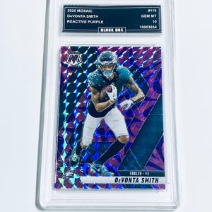 Puede incluir: Una tarjeta de fútbol de De'Vonta Smith de la serie 2025 Mosaic, calificada GEM MT 10, dentro de un soporte protector. La tarjeta presenta un diseño de mosaico con tonos morados y azules, y el texto "EAGLES - #6 De'Vonta Smith".