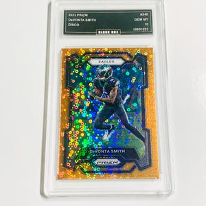 Puede incluir: Una tarjeta coleccionable DeVonta Smith Disco Prizm 2023, calificada GEM MT 10, en un protector. La tarjeta presenta a un jugador con el uniforme de los Eagles con un diseño colorido y la palabra "EAGLES".