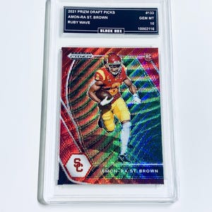 Könnte beinhalten: Eine Football-Sammelkarte mit Amon-Ra St. Brown in roter und goldener Uniform. Die Karte ist in einem transparenten Halter. Auf der Karte stehen "2021 Prizm Draft Picks", "Ruby Wave" und der Name des Spielers.