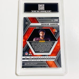Puede incluir: Una tarjeta de f&uacute;tbol calificada con Shedeur Sanders de los Cleveland Browns. La tarjeta tiene un dise&ntilde;o de mosaico con una foto del jugador. La tarjeta est&aacute; encerrada en un soporte protector transparente con el logotipo "Black Box Grading Club".