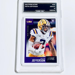 Op de afbeelding: Een beoordeelde 2020 Panini Score Justin Jefferson voetbalkaart. De kaart toont een speler in een wit en paars uniform met een voetbal. De kaart zit in een doorzichtige beschermhoes met de tekst "2020 PANINI SCORE" en "JUSTIN JEFFERSON".