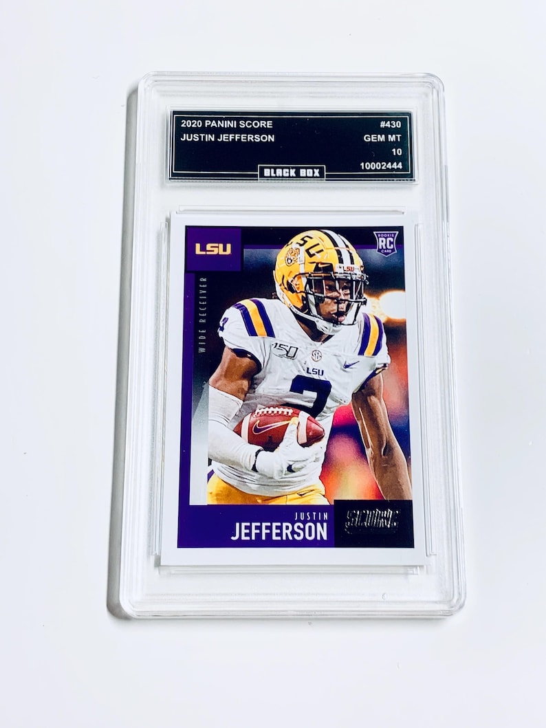 Op de afbeelding: Een beoordeelde 2020 Panini Score Justin Jefferson voetbalkaart. De kaart toont een speler in een wit en paars uniform met een voetbal. De kaart zit in een doorzichtige beschermhoes met de tekst "2020 PANINI SCORE" en "JUSTIN JEFFERSON".